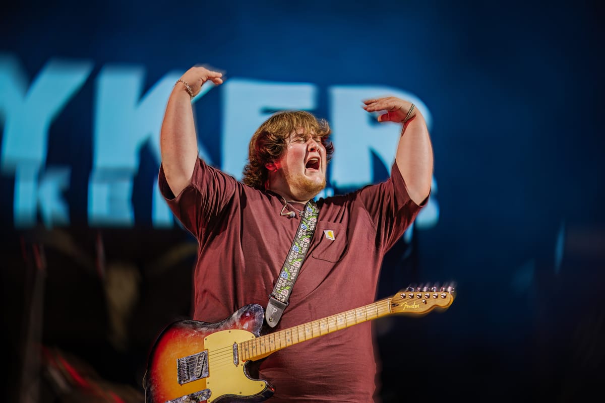 Guitarist mid-song with both arms raised over his head against a blue backdrop