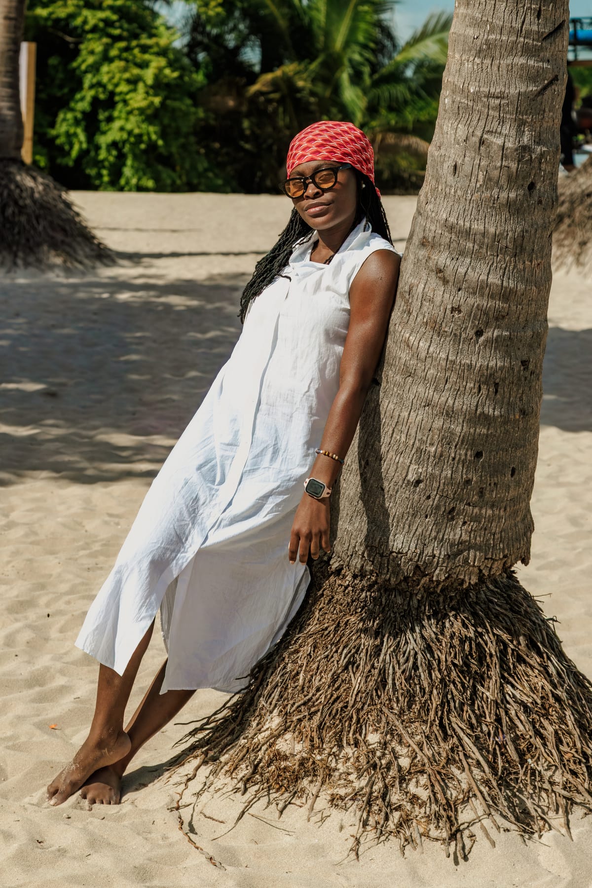 Portrait of a woman in a white dress and red headwrap leaning against a palm tree on a sandy beach