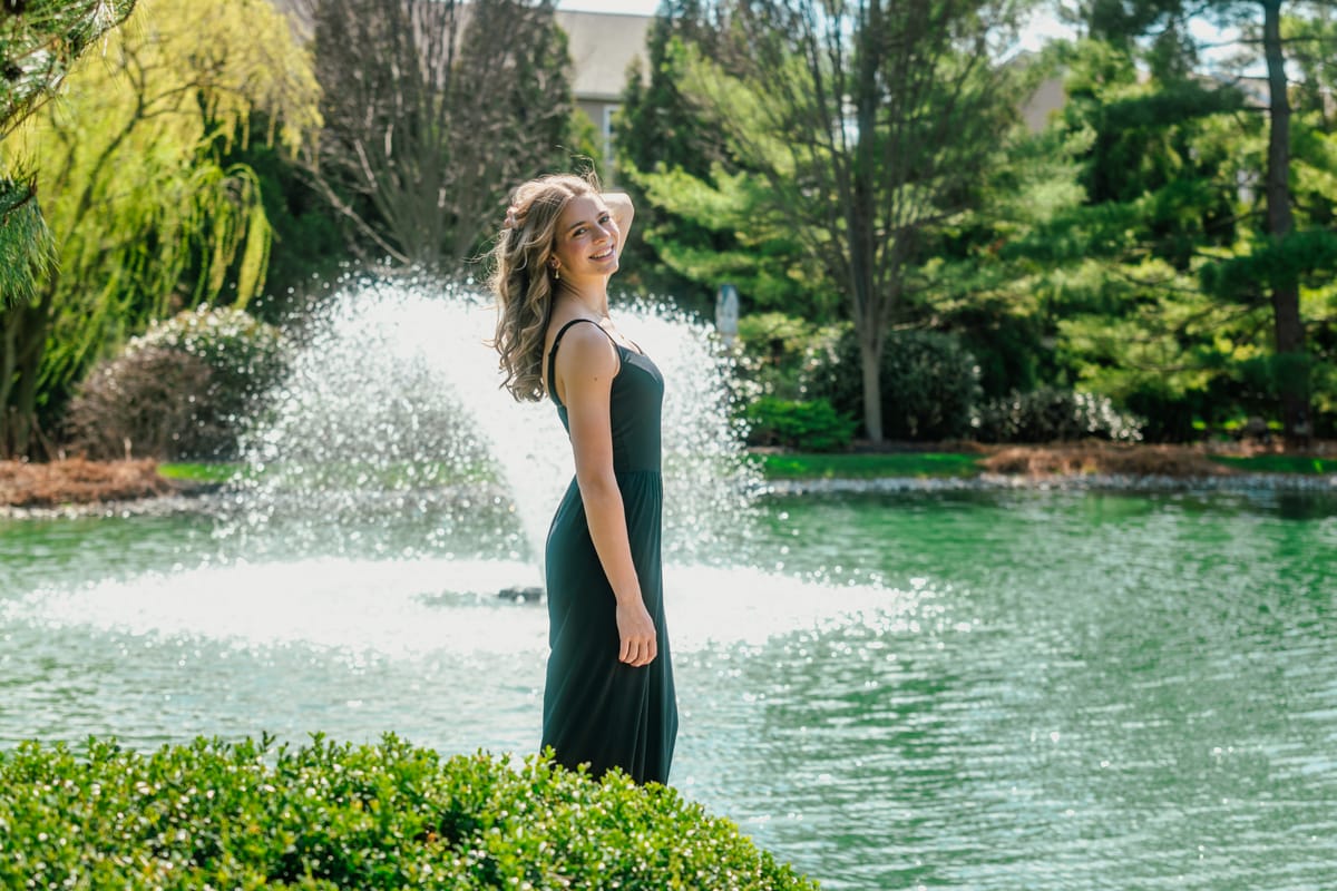 Senior portrait of a woman in a green dress beside a garden pond with a fountain behind her