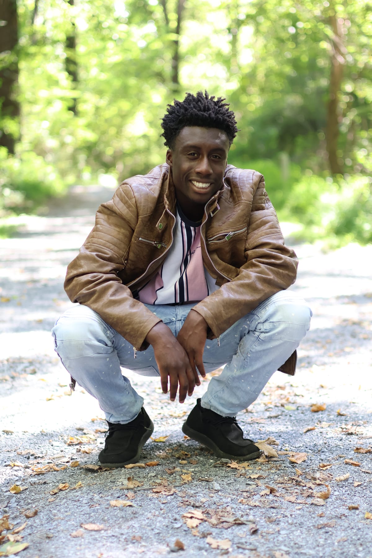 Portrait of a man in a tan leather jacket crouched on a wooded path, smiling