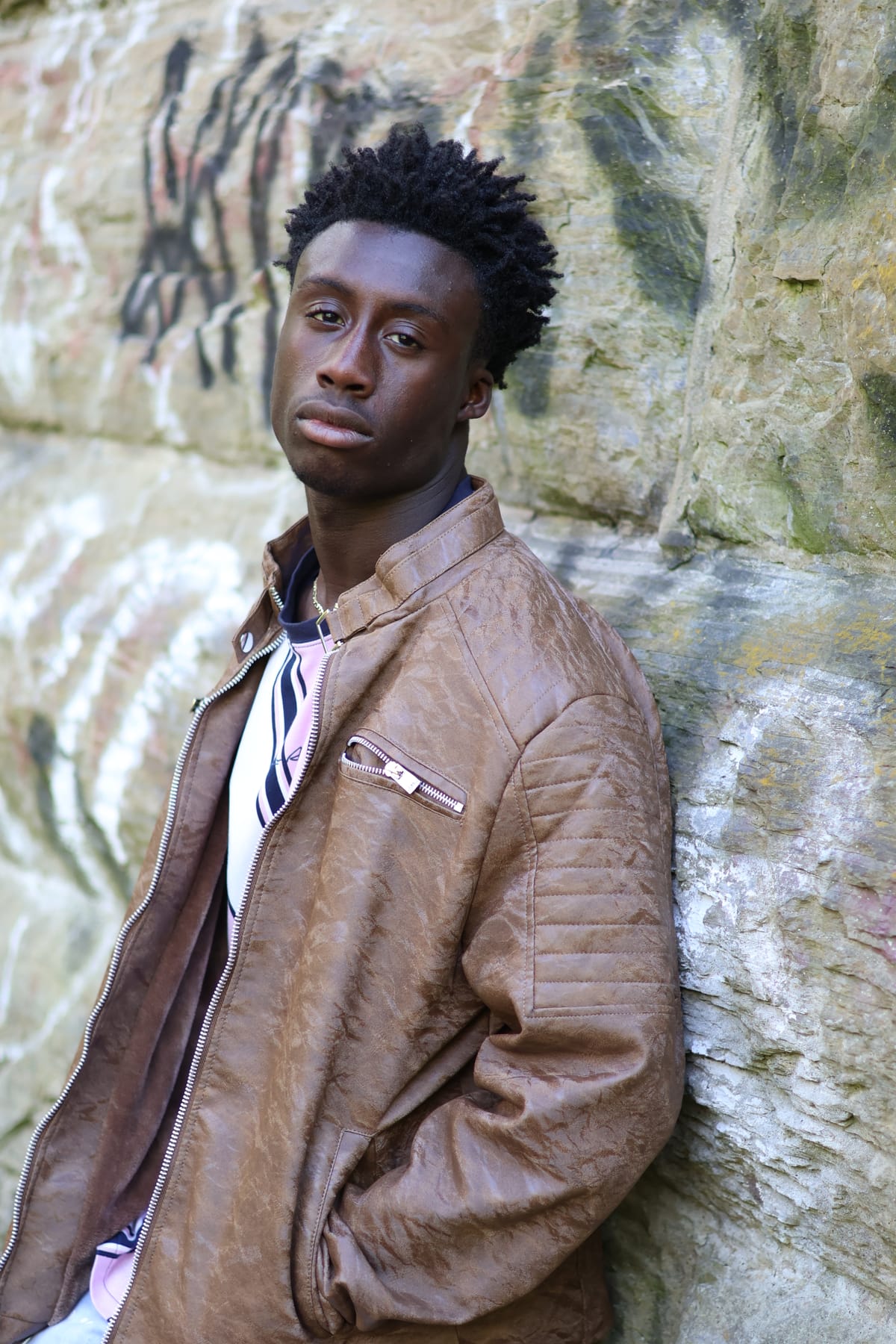 Close portrait of a man in a tan leather jacket resting against a moss-streaked rock