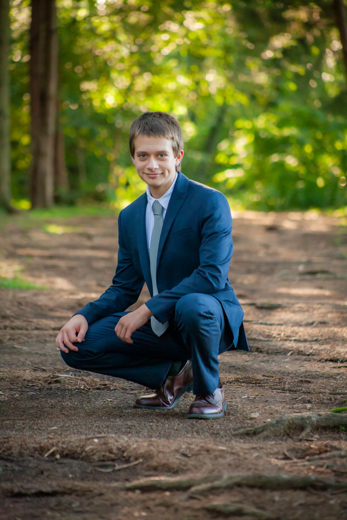 Senior portrait of a young man in a navy suit crouched on a woodland path