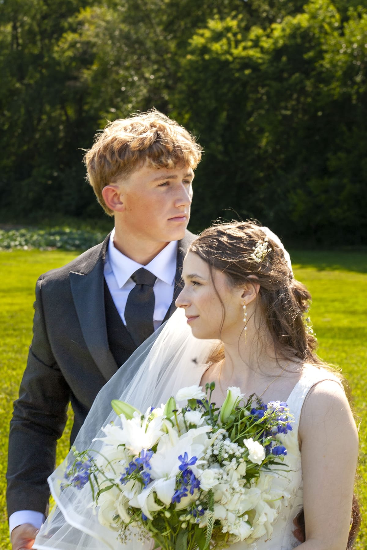 Groom and bride standing close on a sunlit lawn, veil catching the breeze